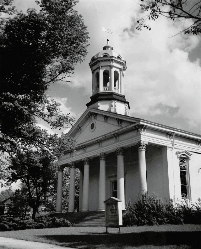 An unidentified church building in the neoclassical style with a view of the front doors and steeple. The church sign is visible in front of the building, but illegible.